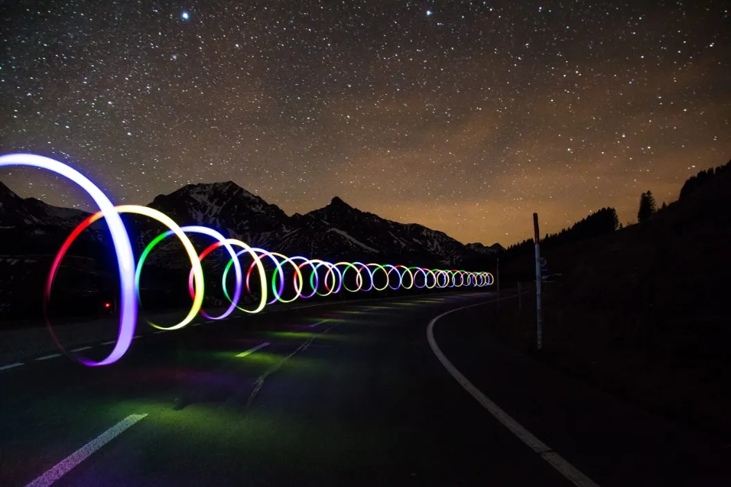 Long-exposure photo of colorful light trails on a road under a starry night sky with mountains in the background.