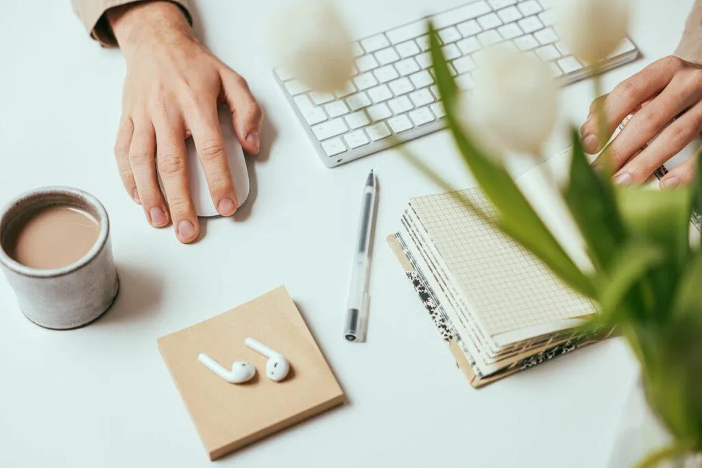 A person has one hand on a mouse and the other on a gridded notepad, on a desk holding coffee and flowers.
