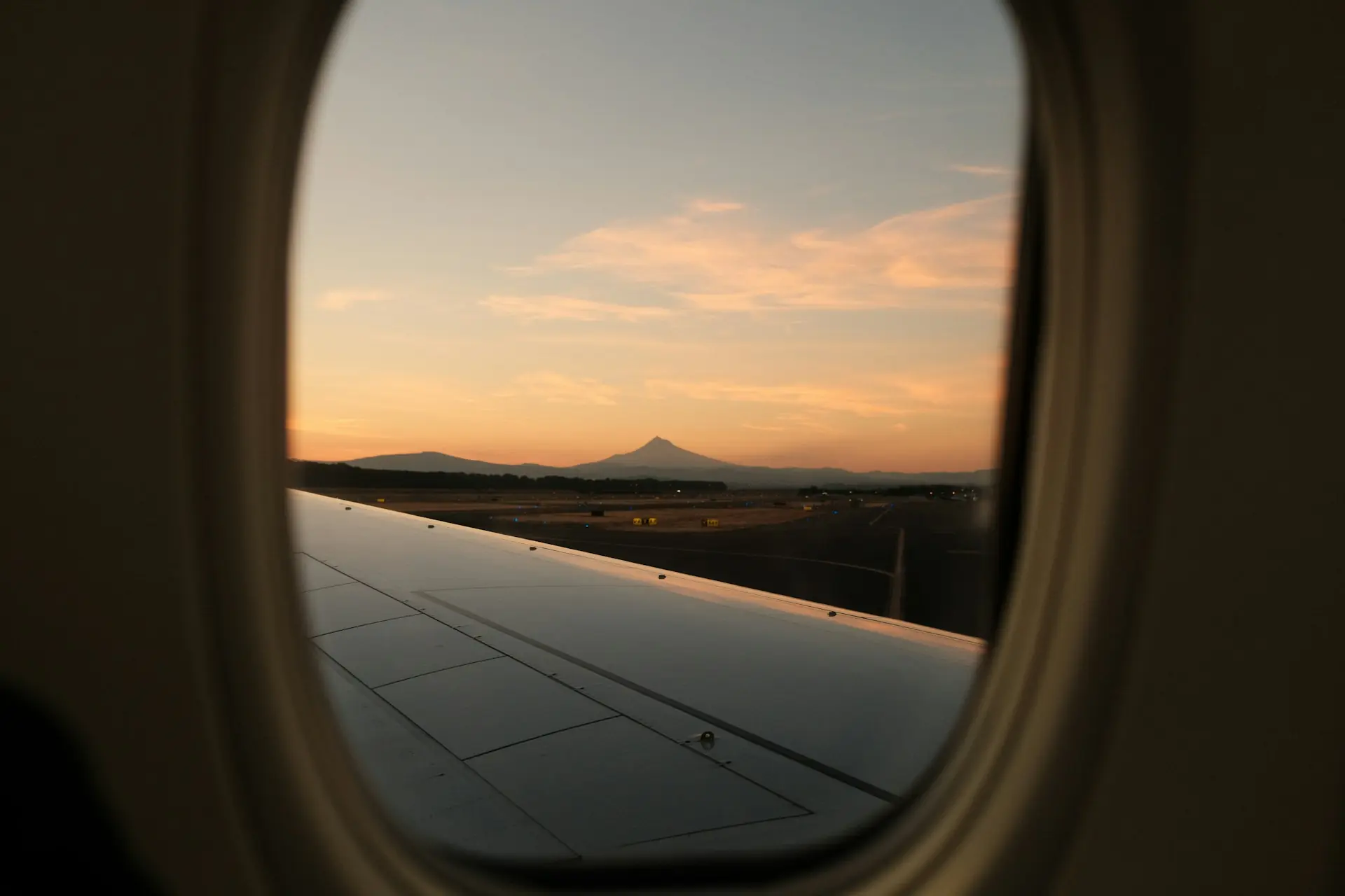A serene sunset view from an airplane window, showing a wing and a distant mountain with a runway in the foreground.
